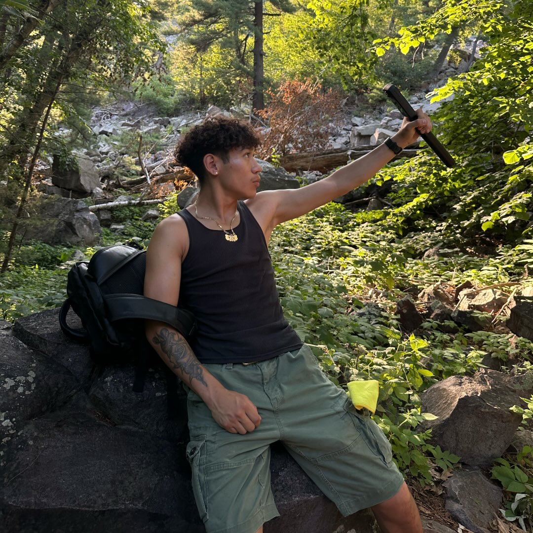 Portrait of Jose Calderon sitting on a rock in a forest, holding a camera rig